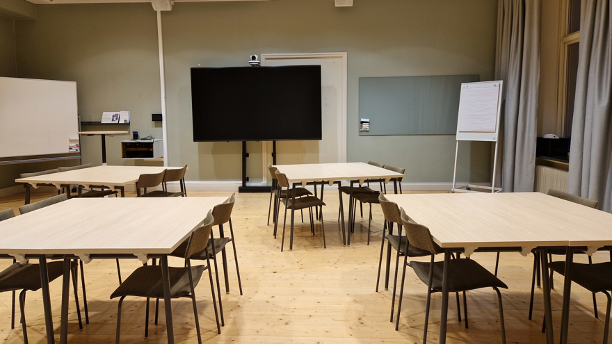 Bord möblerat i grupper, väggfast whiteboardtavla samt en större på hjul och en bildskärm på vänster sida av lokalen.