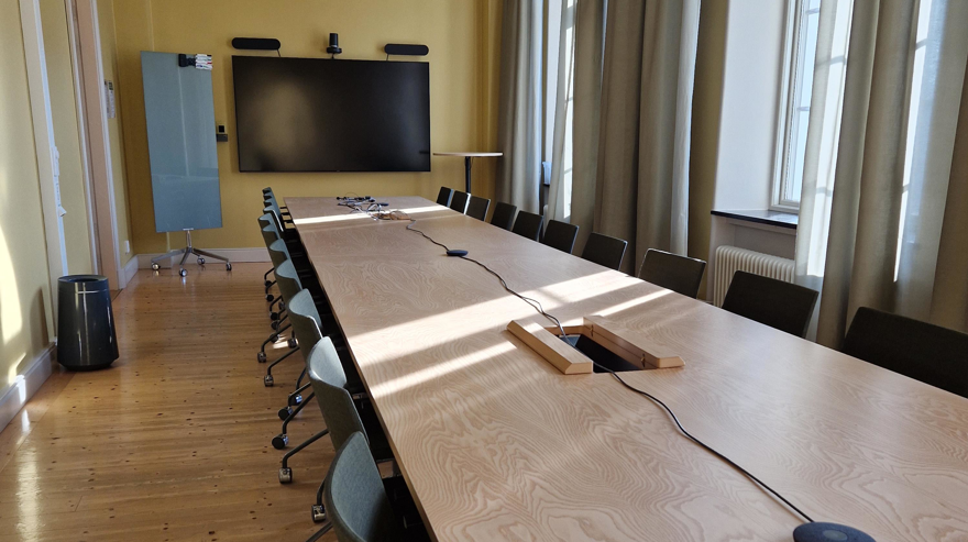 Avlångt bord och stolsfåtöljer på hjul, bildskäm på kortsidan samt en avlång whiteboardtavla på hjul.