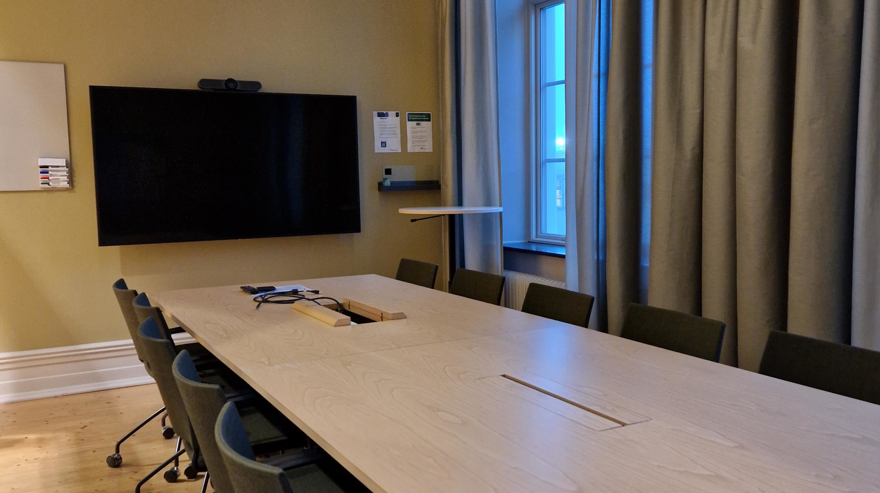 Avlångs bord med stolar på hjul samt bildskärm och liten whiteboardtavla på kortsidan av lokalen.
