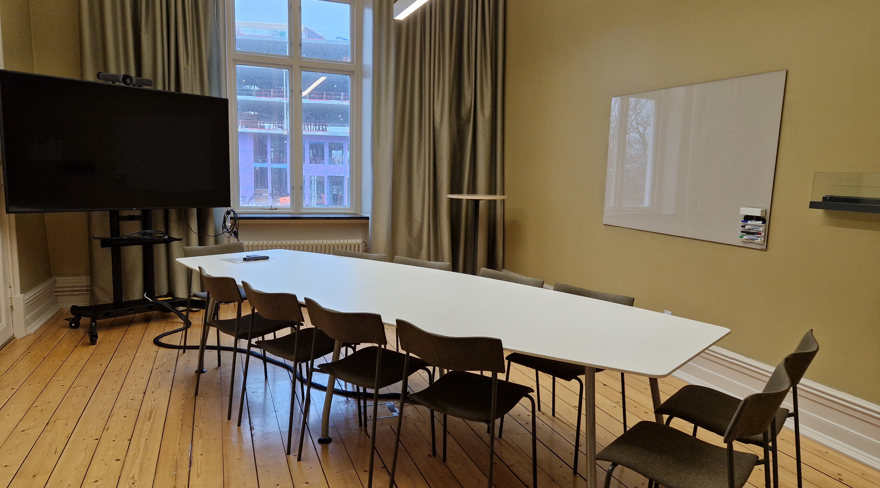 Avlångt konferensbord med stolar samt väggfast whiteboardtavla på höger sida och bildskärm i hörnet av lokalen.