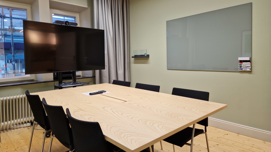 Konferensbord med stolar samt väggfast whiteboardtavla på höger sida och en bildskärm på kortsidan av lokalen.