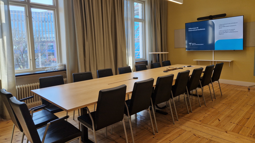 Avlångt bord med stolar samt bildskärm och whiteboardtavla på kortsidan av lokalen.