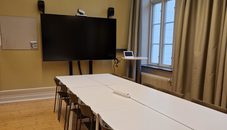 Avlångt bord md stolar samt bildskärm och liten whiteboardtavla på kortsidan av rummet.